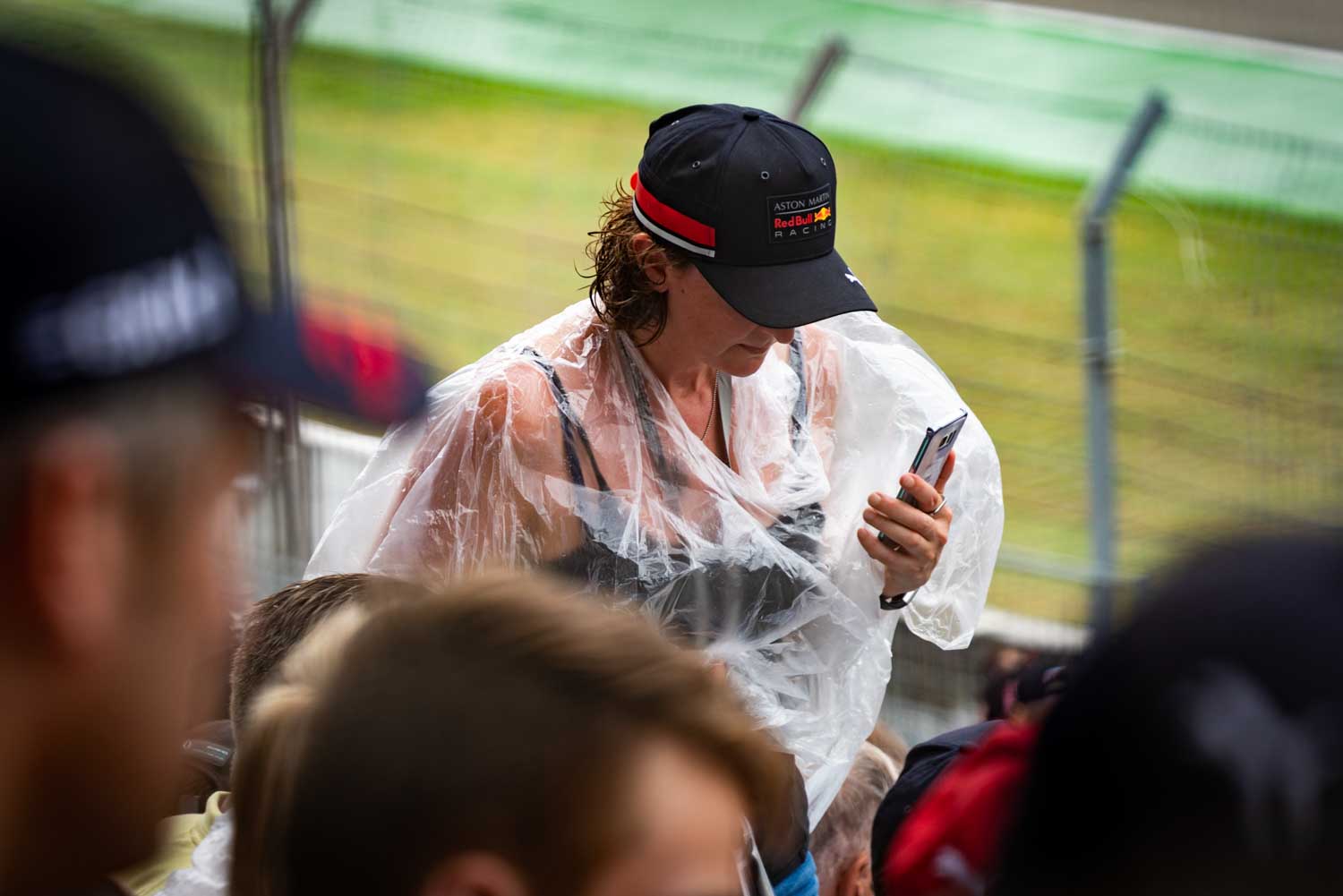 Person in rain poncho and cap using smartphone at an outdoor event with blurred crowd in the foreground.