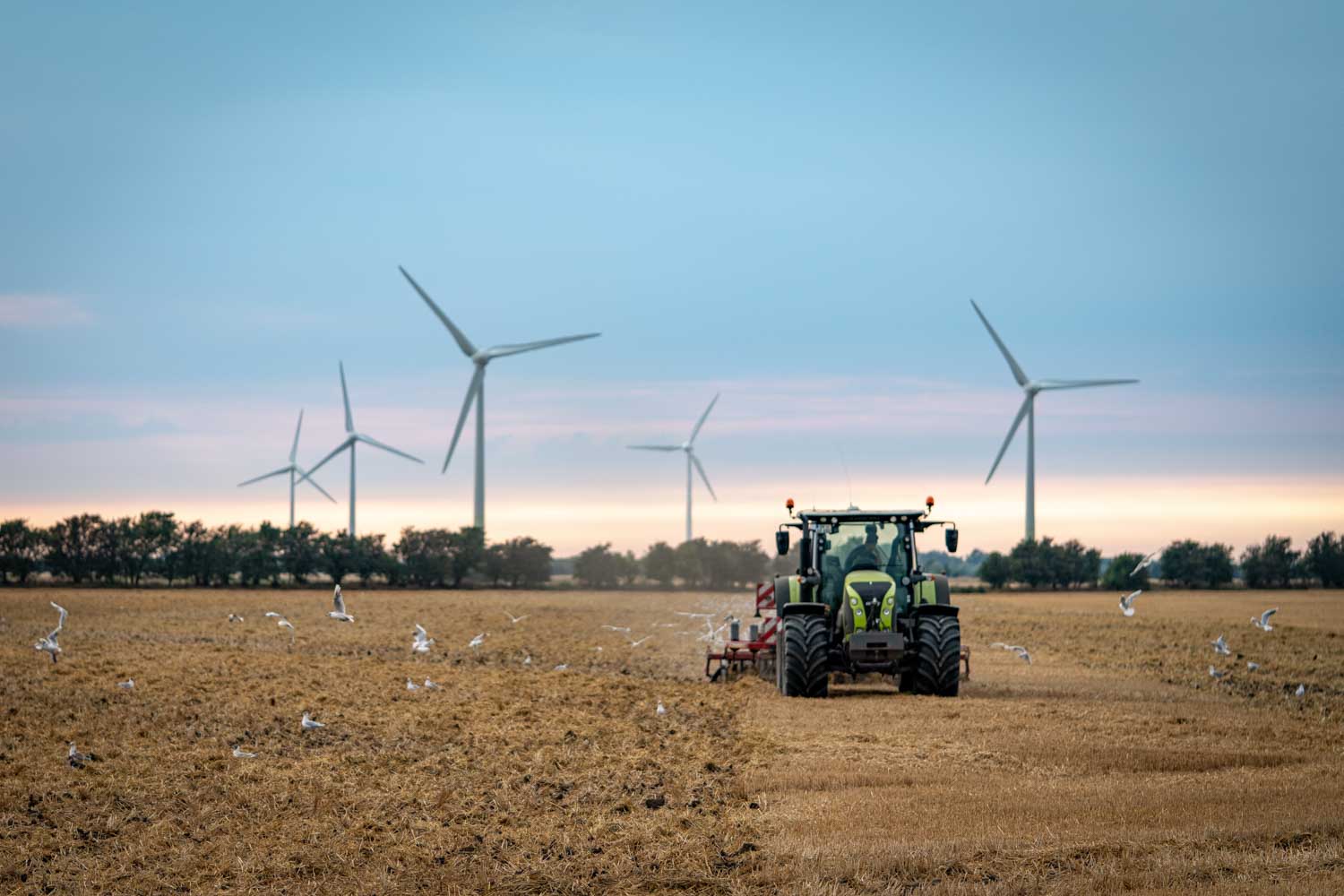 Green tractor plowing a field with wind turbines in the background, under a clear sky.