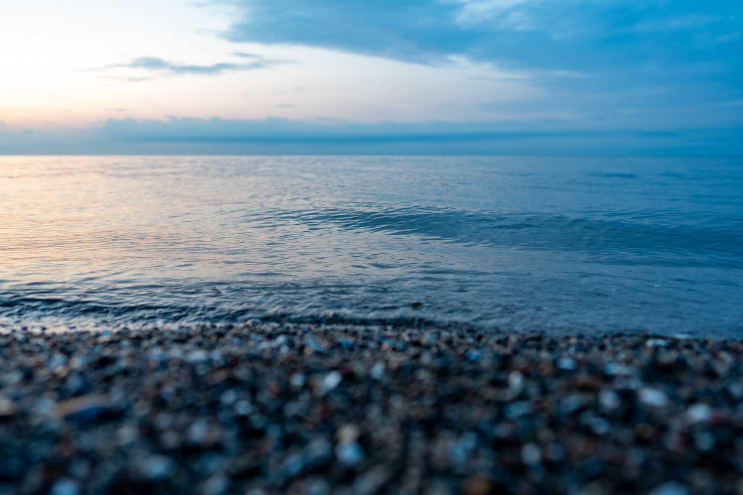 Calm sea waves gently lapping against a pebbly shore at sunrise under a partly cloudy sky.