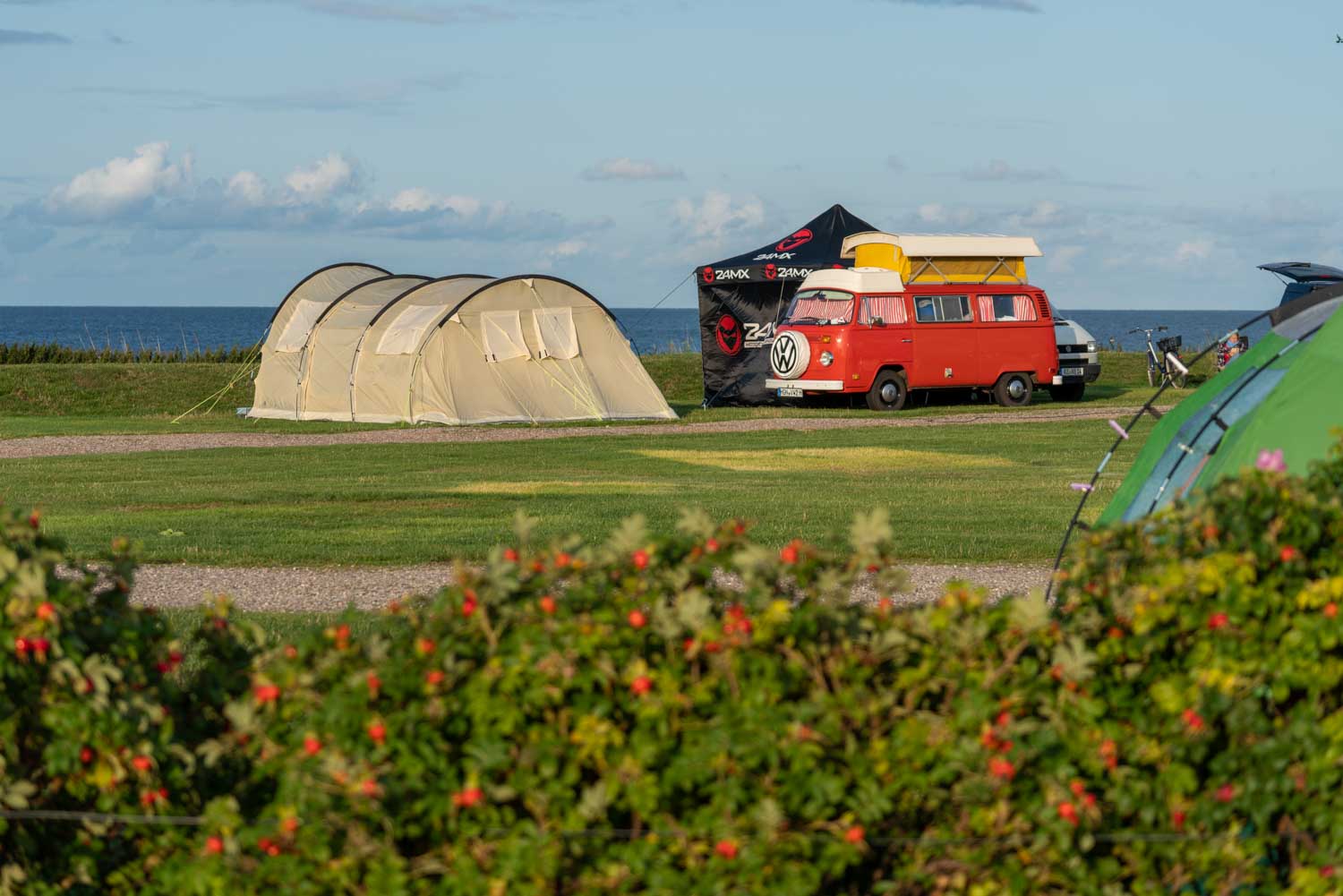 Vintage red camper van and tents on a grassy seaside campsite under blue skies.