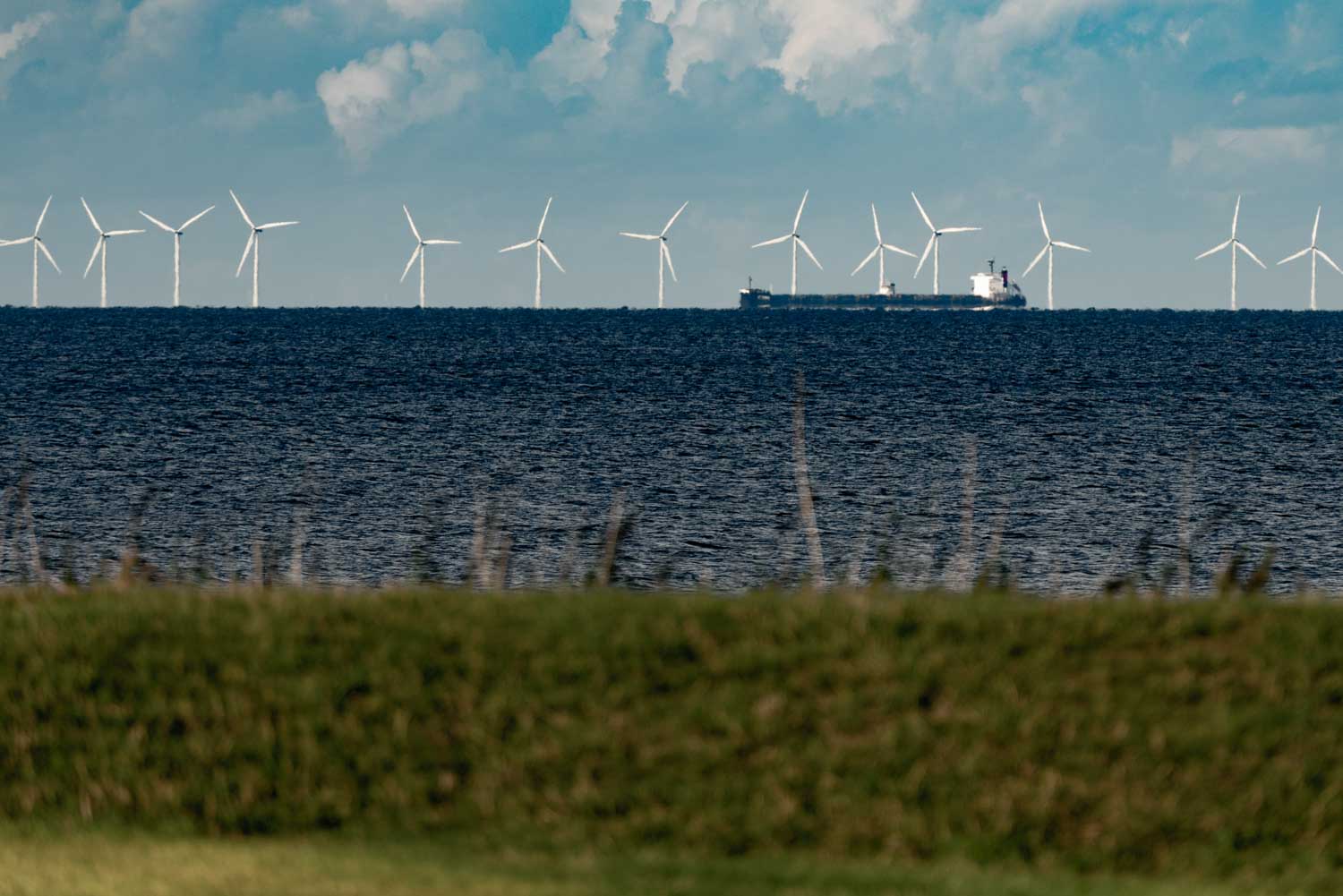 Distant wind turbines on a calm sea with a ship passing, viewed from a grassy shore under a cloudy sky.