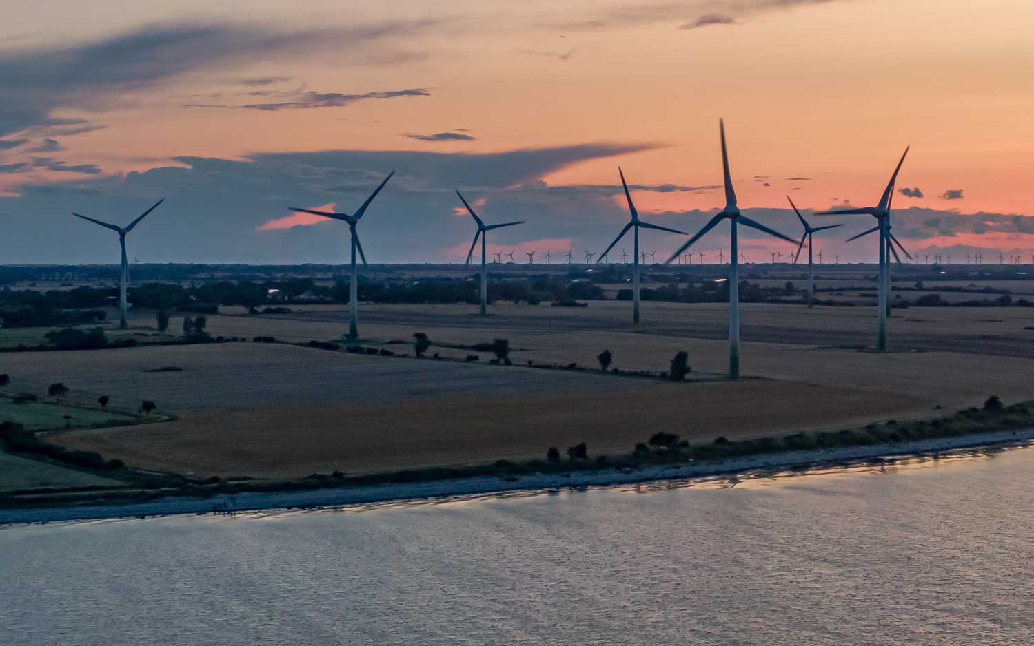 Wind turbines on farmland at sunset, near a calm river, under a pink and blue sky.
