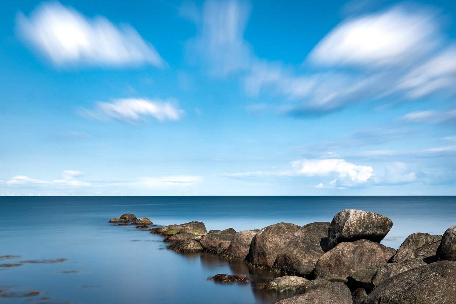 Smooth ocean scene with large rocks and a vibrant blue sky dotted with fluffy clouds.