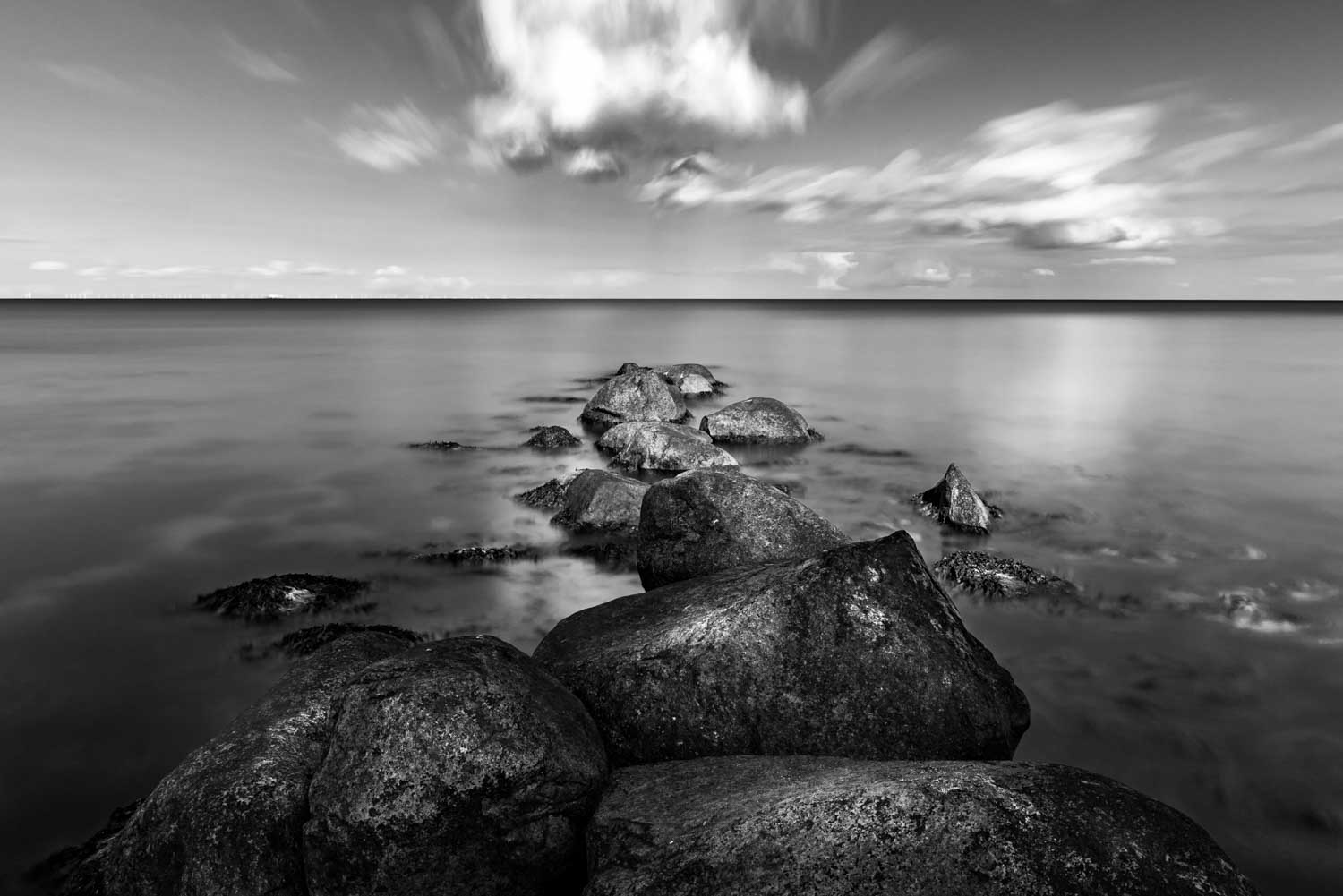Black and white seascape with rocks leading into calm ocean under a cloudy sky.