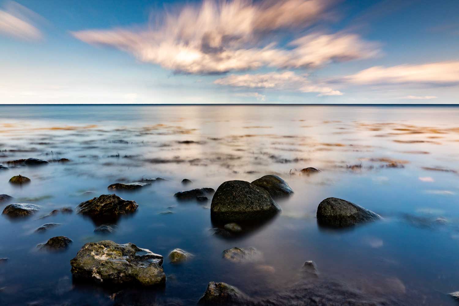 Tranquil seascape with smooth stones in water, blurred clouds in blue sky reflecting on ocean at sunset.