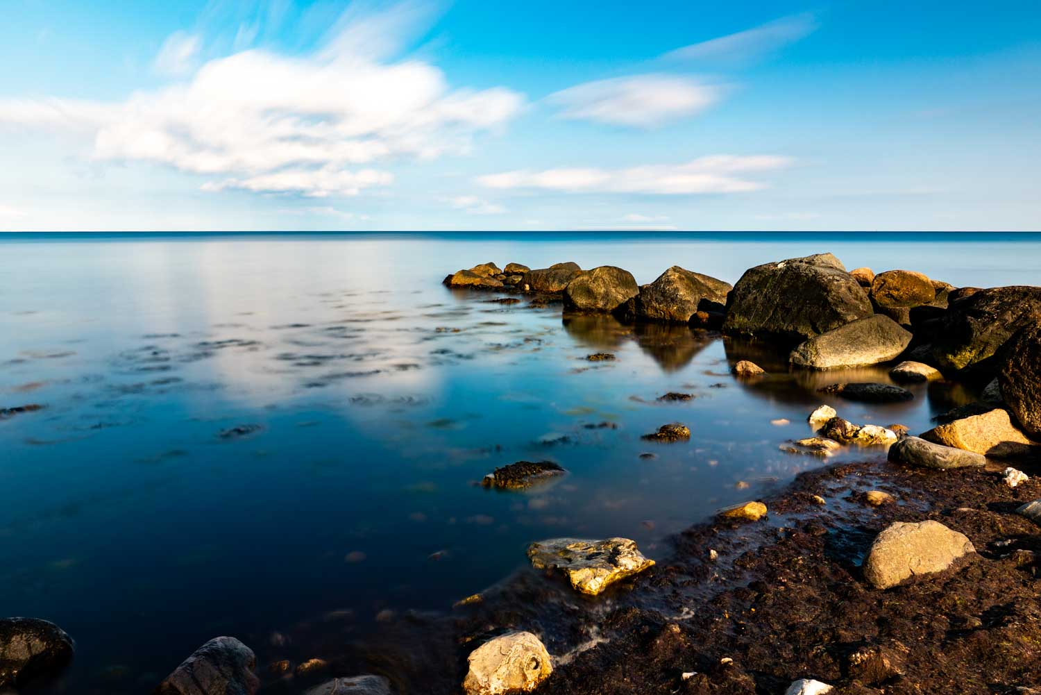 Calm sea with rocks under a blue sky and fluffy clouds reflecting on water.