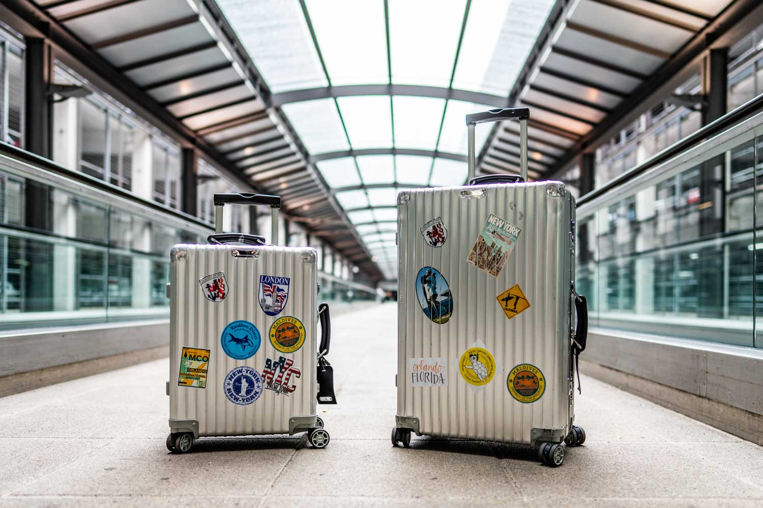 Two sticker-covered silver suitcases stand on an airport walkway under a glass roof.