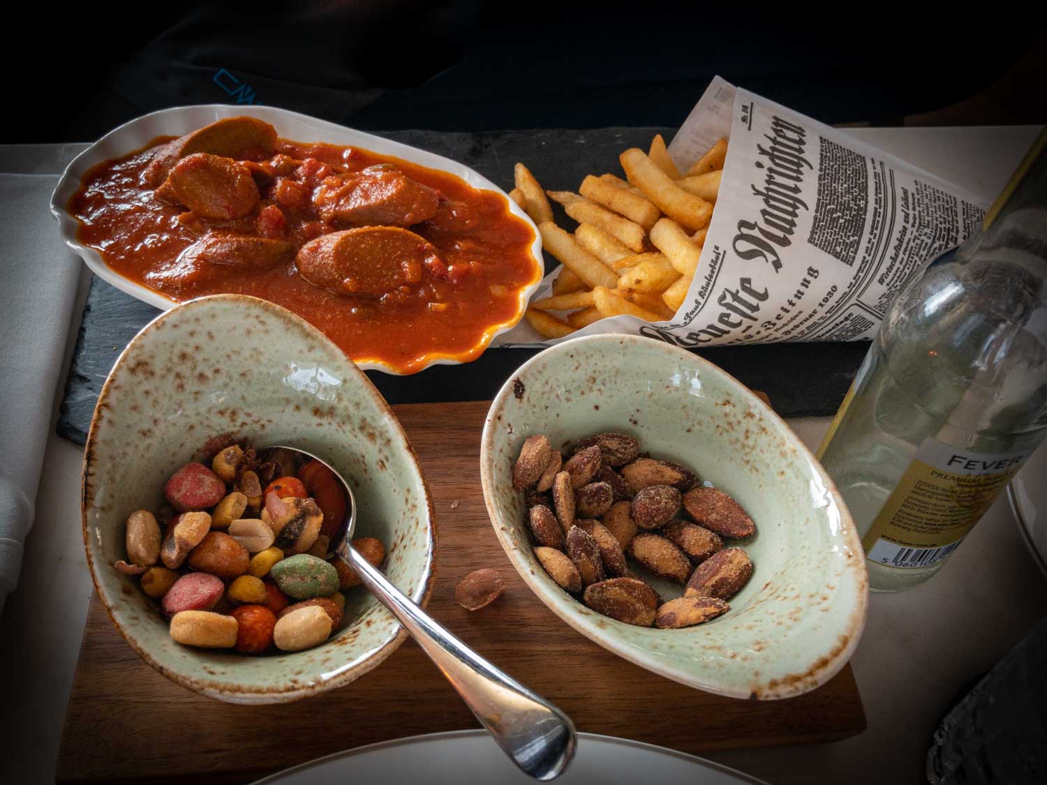 Spicy sausage in tomato sauce with fries, mixed nuts, and almonds in bowls, alongside a Fever-Tree mixer bottle.