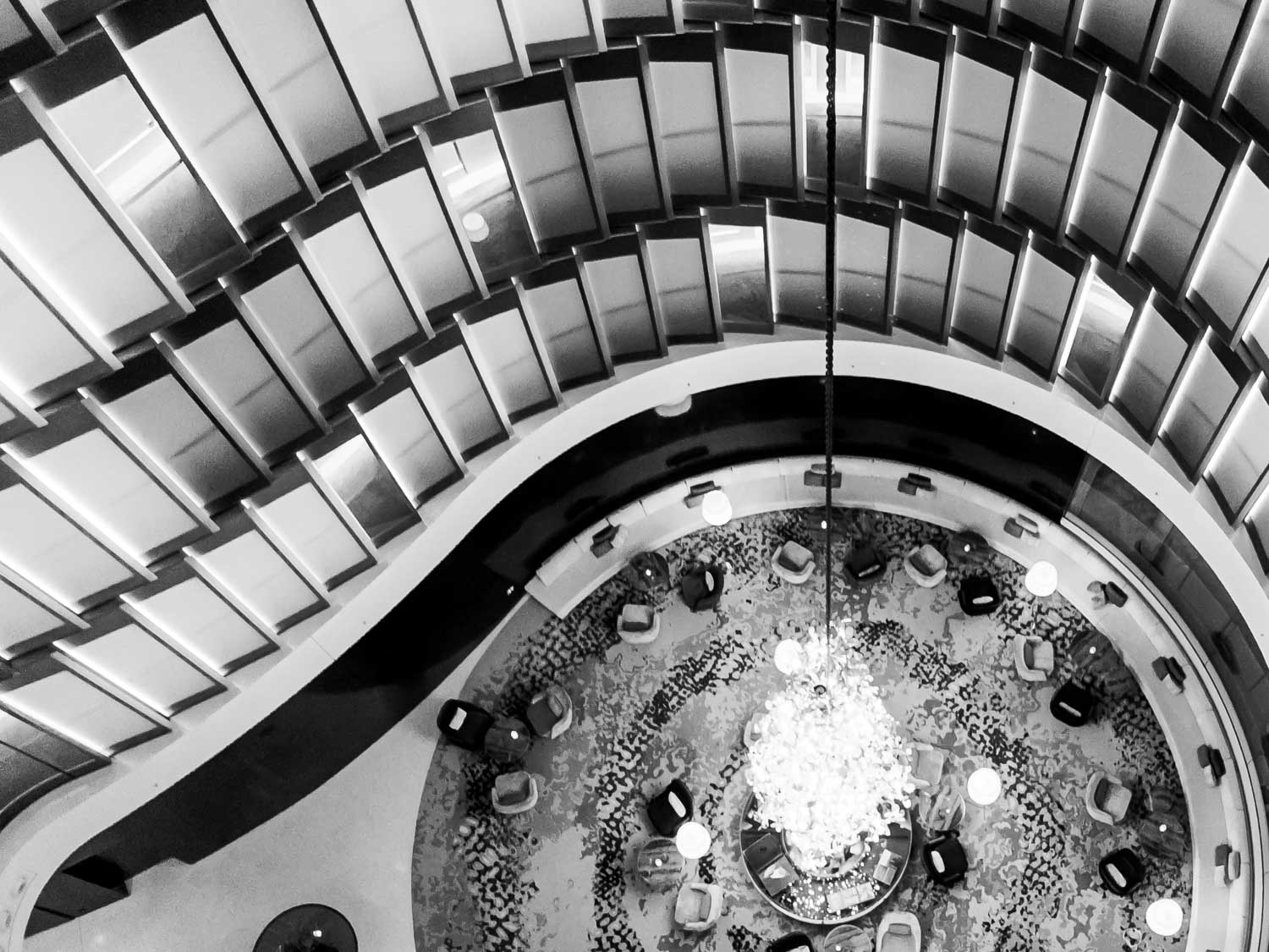 Stylish modern atrium with curved balcony and large chandelier, overlooking patterned seating area from above.