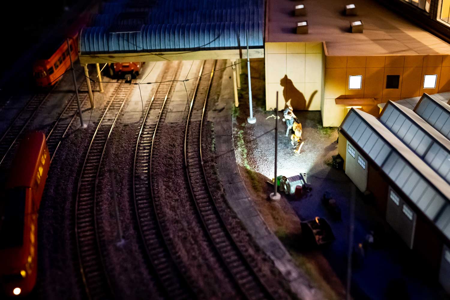 Model railway at night with trackside building and dinosaur figures casting shadows under artificial light.