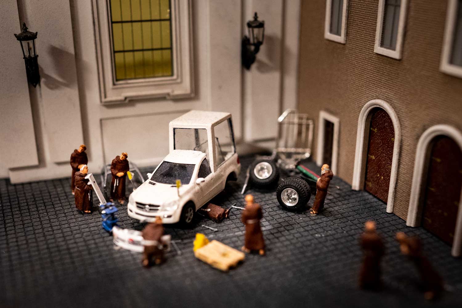 Miniature scene of robed figures fixing a white car in a courtyard with spare tires on the ground.