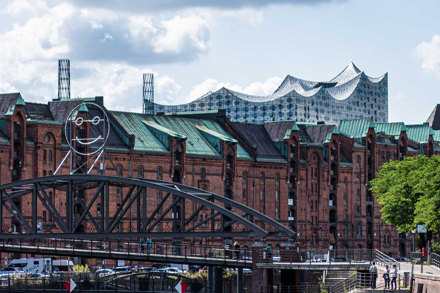 Historic brick warehouses with modern Elbphilharmonie in Hamburg skyline under cloudy sky, featuring a smiling face sculpture.