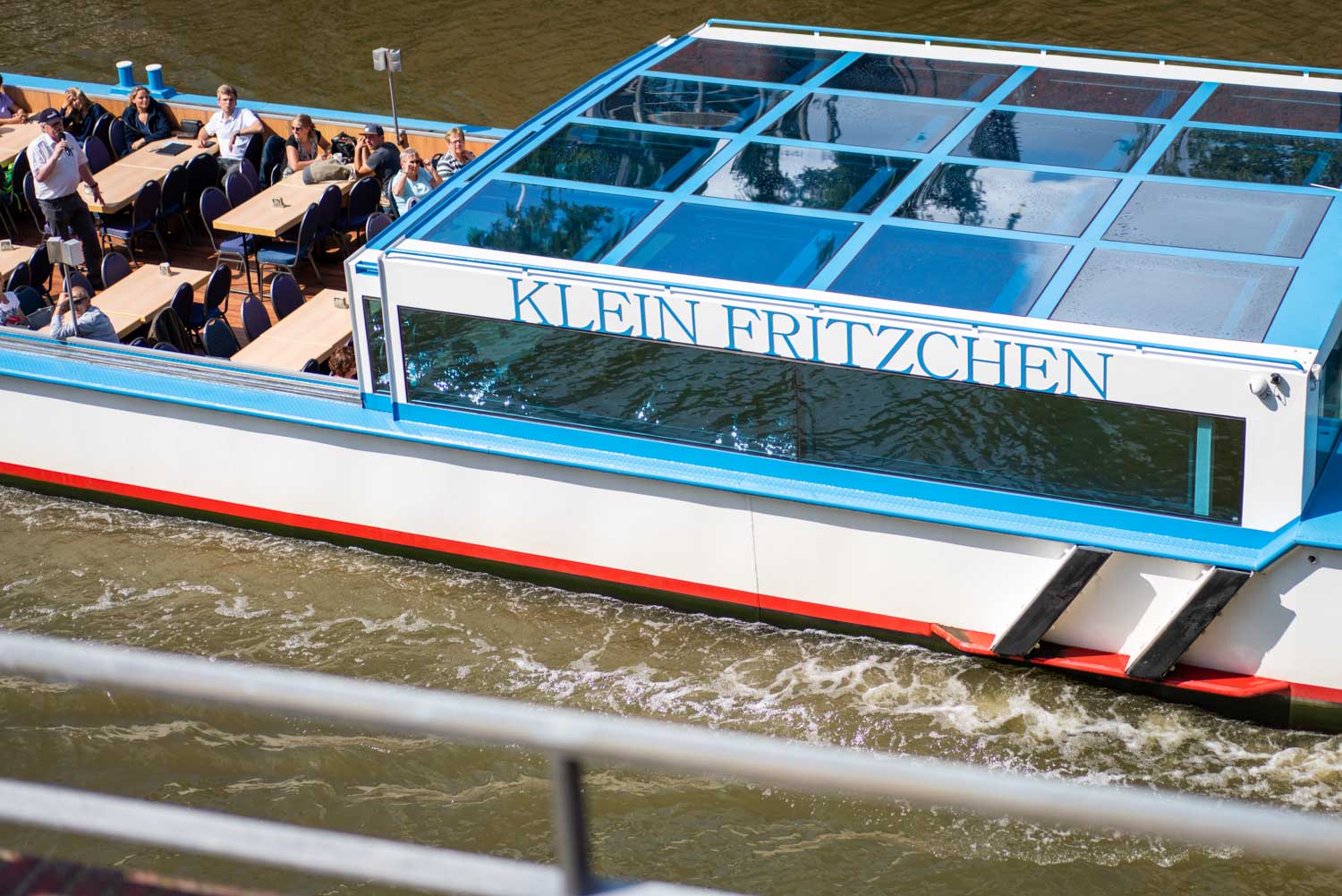 Tour boat Klein Fritzchen cruises a river with passengers under a sunny sky, featuring a glass roof and outdoor seating.