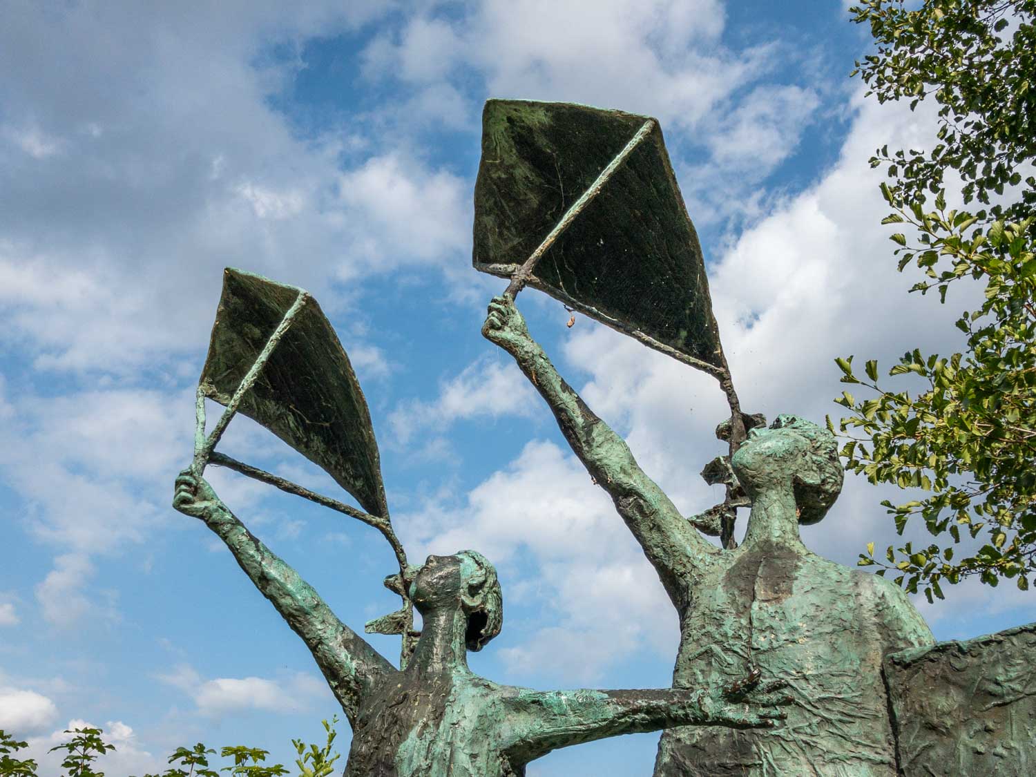Bronze statue of two figures flying kites against a blue sky with scattered clouds and surrounding greenery.