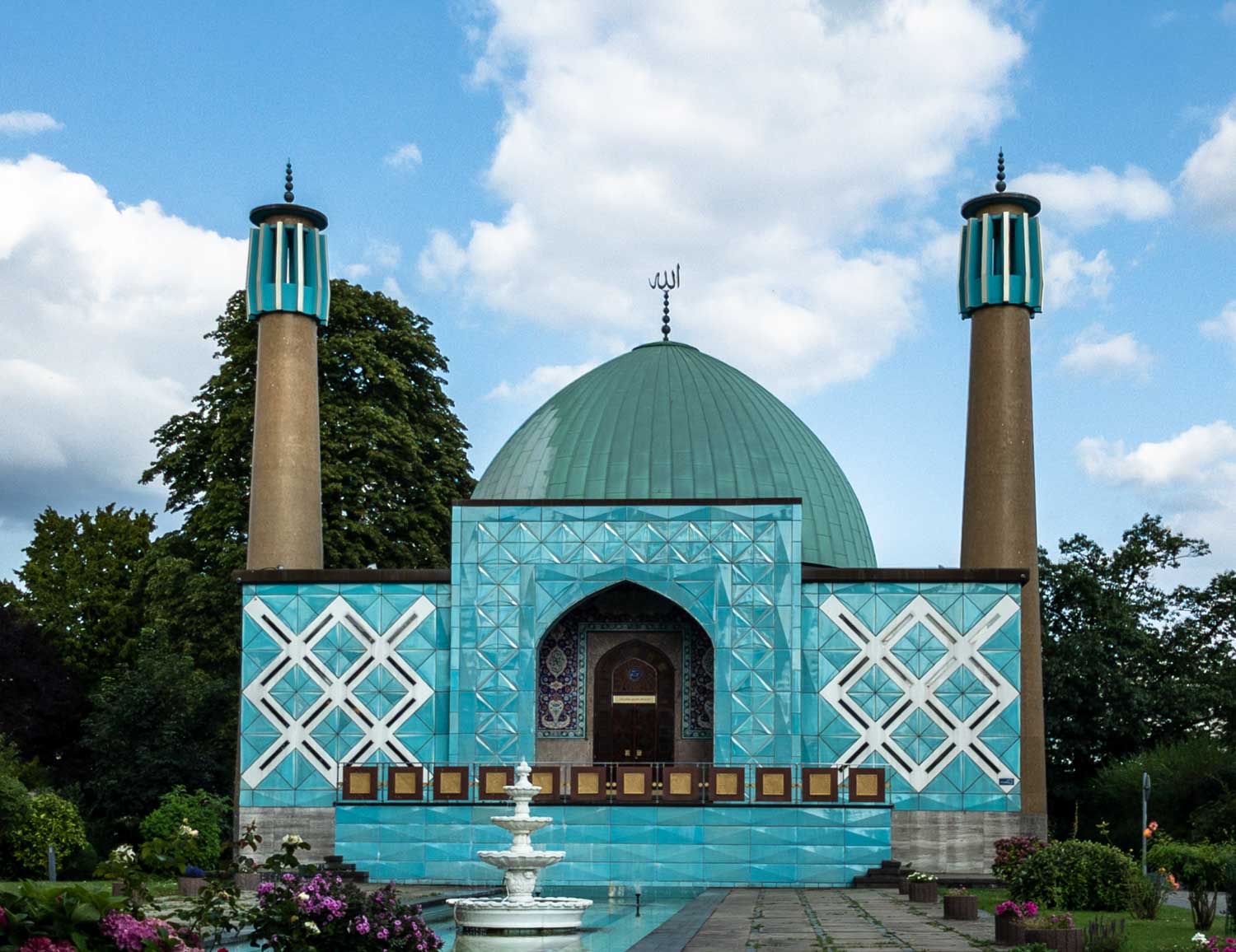 Turquoise mosque with geometric patterns, twin minarets, and a fountain in a lush garden setting under a cloudy blue sky.
