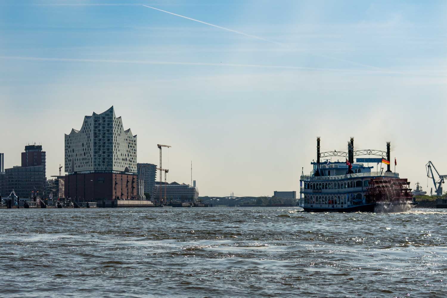 Paddle steamer on Elbe River with Elbphilharmonie and Hamburg skyline in the background under a clear blue sky.