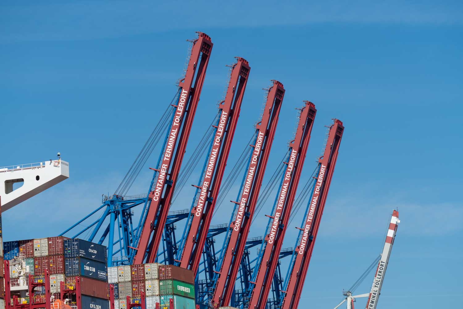 Red shipping cranes at Container Terminal Tollerort, handling stacked cargo containers under a clear blue sky.