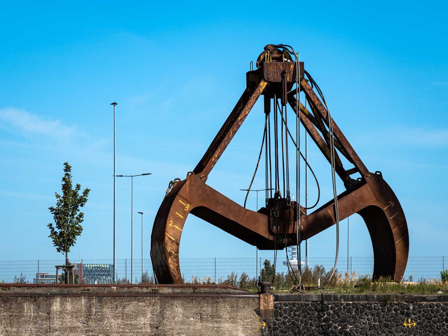 Rusty industrial grab crane under a blue sky with a small tree beside it.