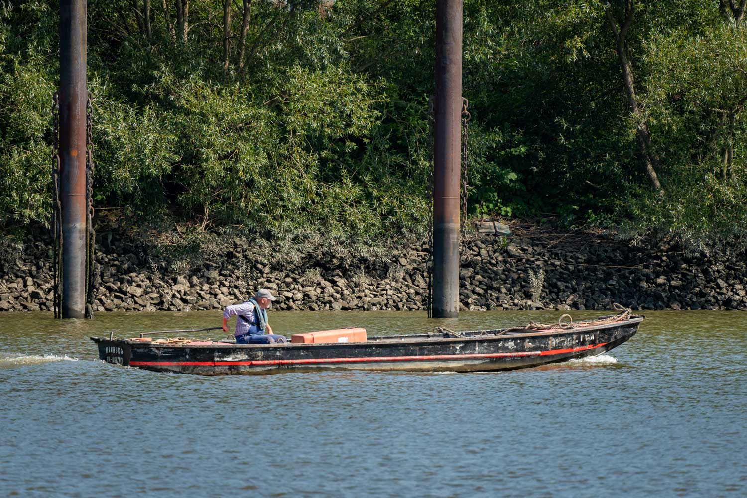 Man in a boat navigating a river by lush greenery and metal poles, clear sunny day.