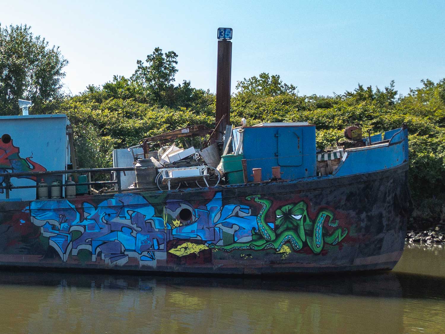 Graffiti-covered ship with colorful art, docked in a calm river, surrounded by lush greenery.