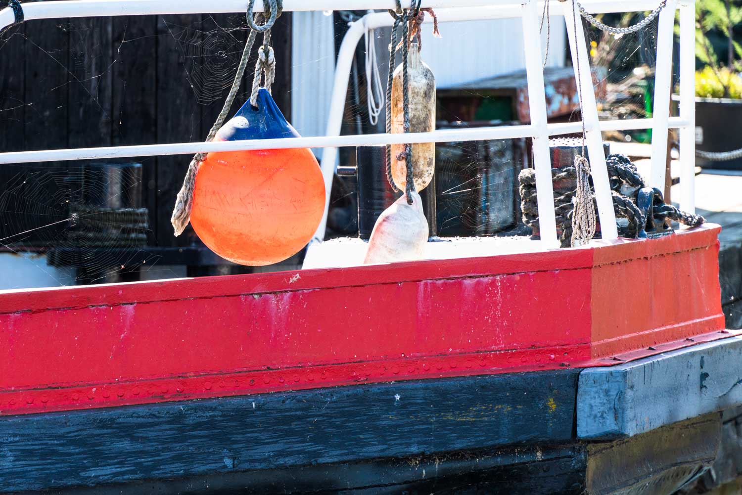 Orange and white buoys hanging on a red and black fishing boat railing with visible spiderwebs.
