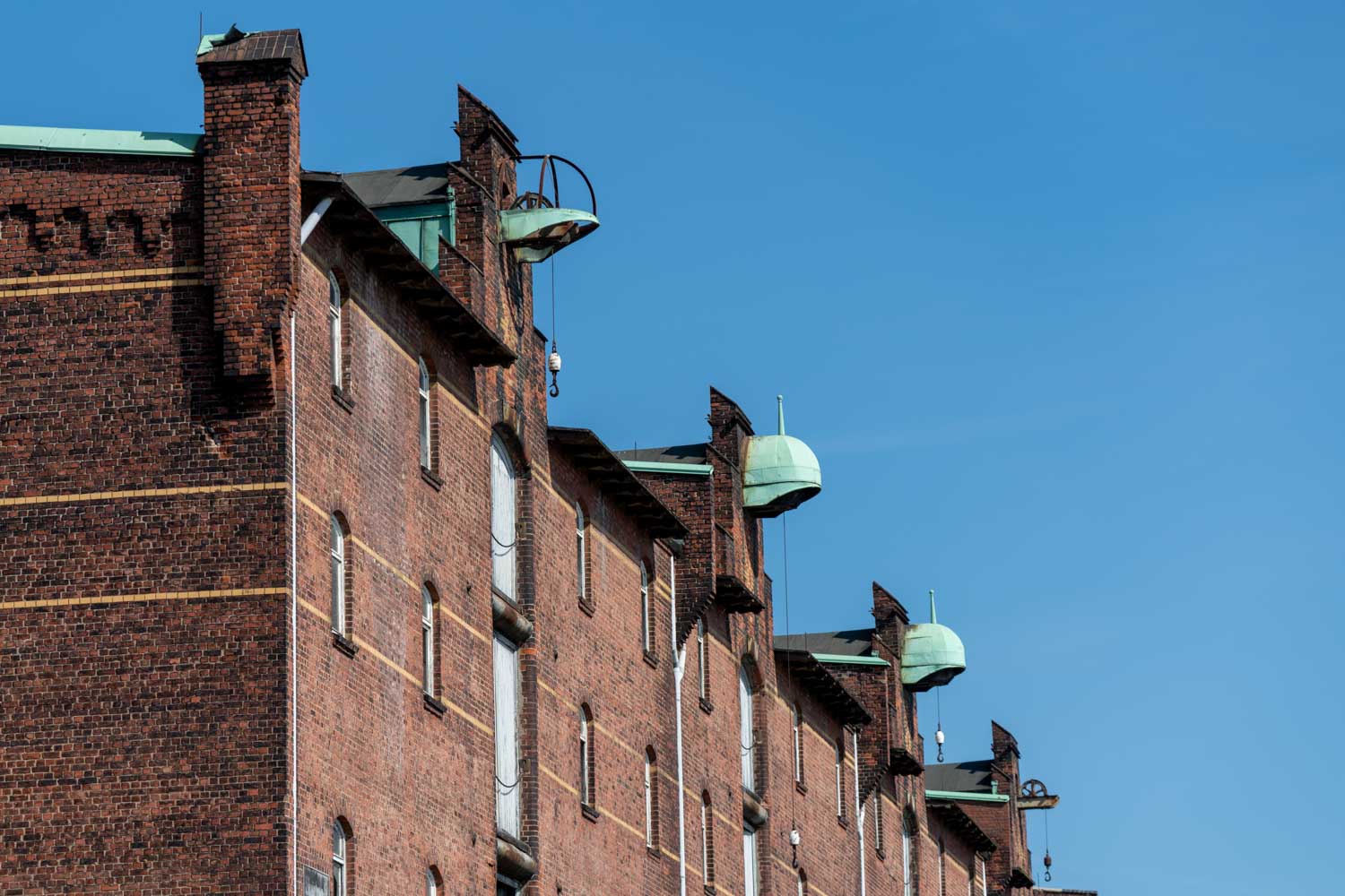 Historic brick warehouse with green domed roofs and hooks against a clear blue sky.
