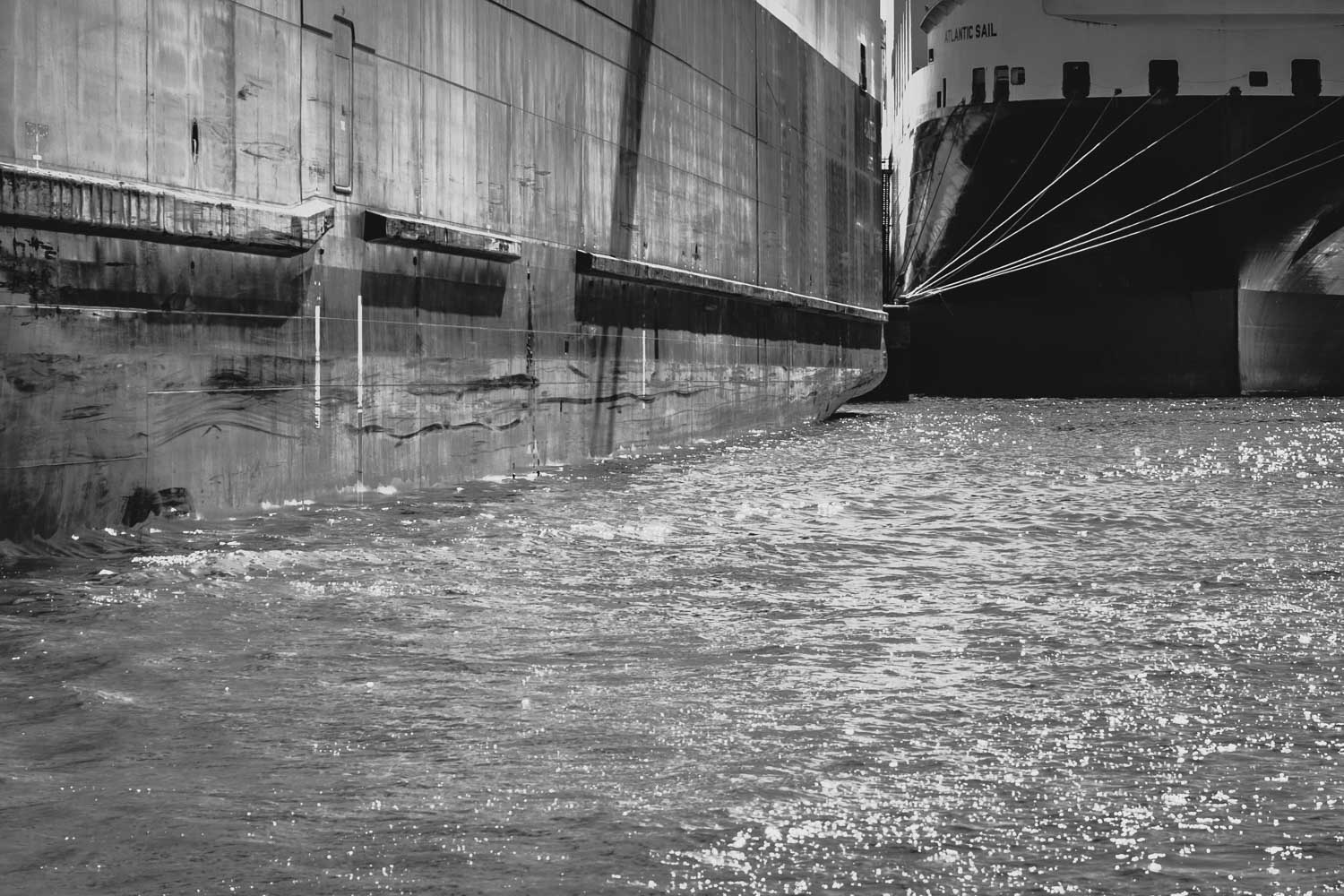 Black and white photo of large ships docked side by side, with sunlight reflecting on the water surface.
