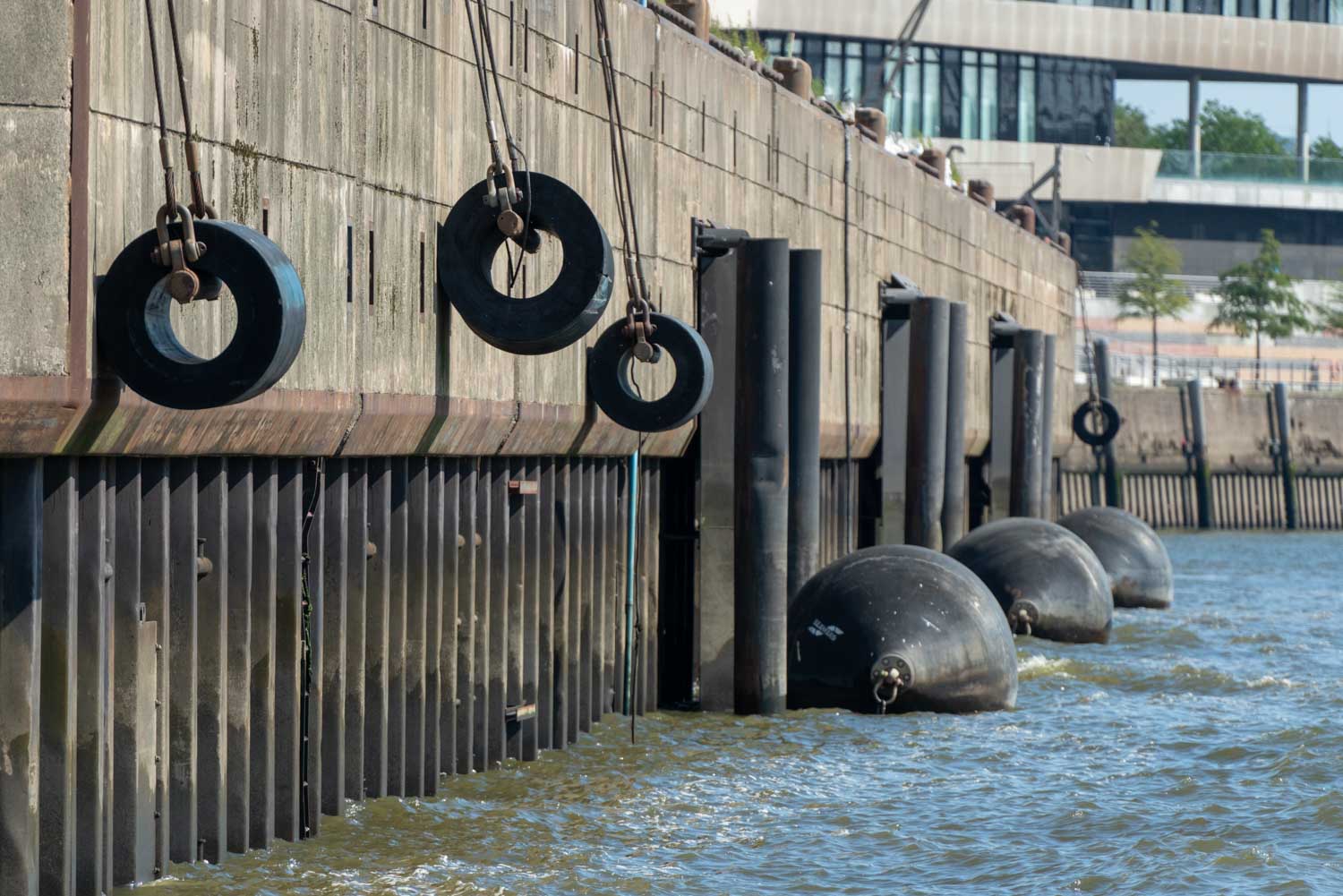 Dock with hanging tires and round bumpers on water, adjacent to a concrete wall with modern building in background.