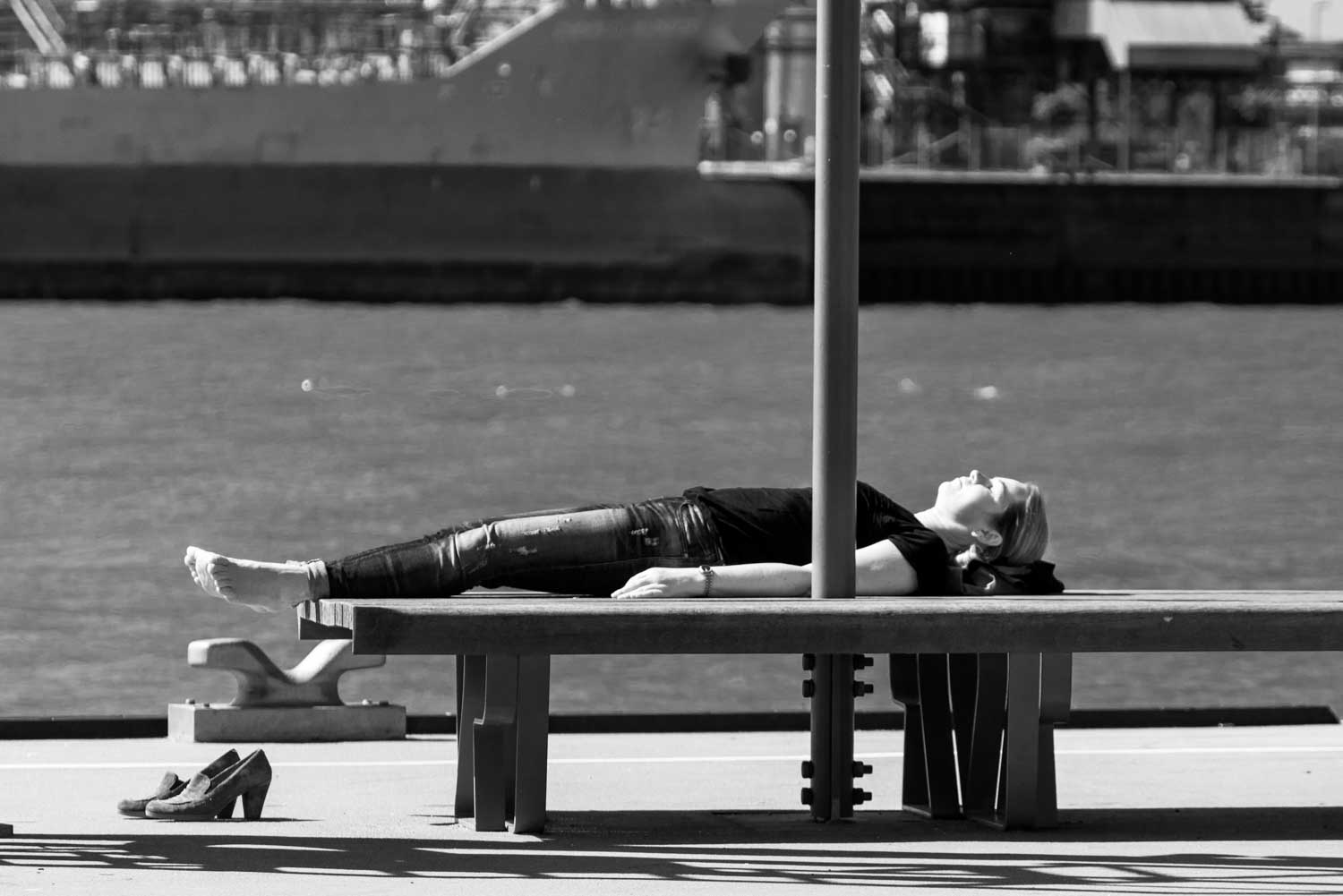 Woman relaxing barefoot on a bench by the waterfront, with shoes beside her, enjoying a sunny day.