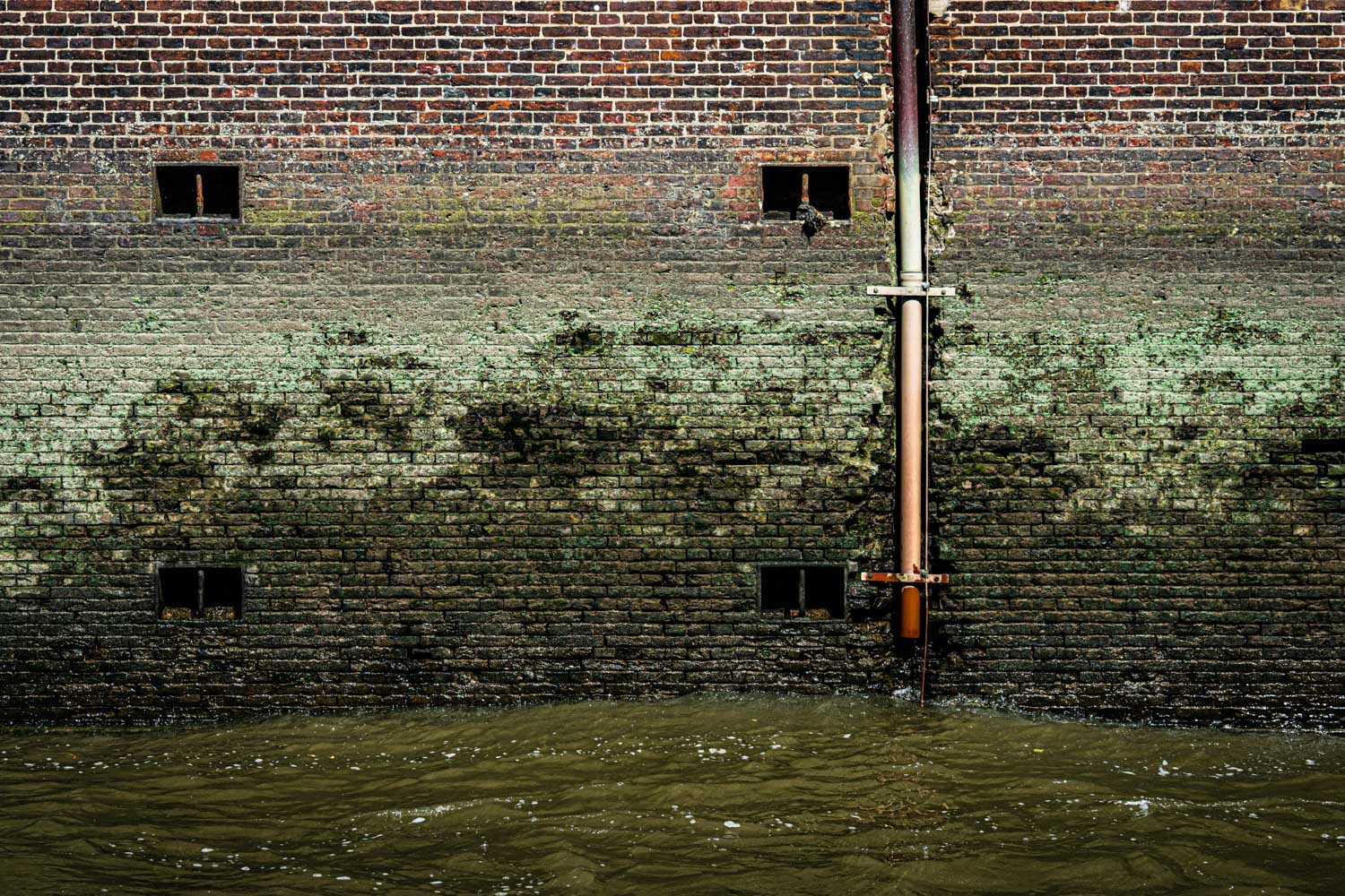Moss-covered brick wall with drainage pipe and small windows above water.