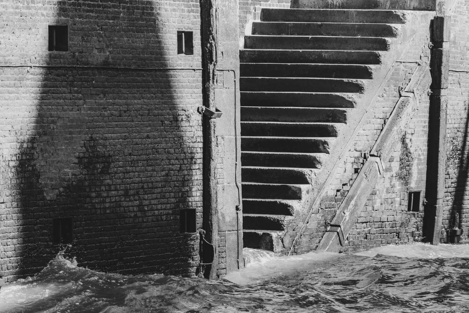 Black and white image of stone steps beside a brick wall with water at the base, creating a dramatic shadow contrast.