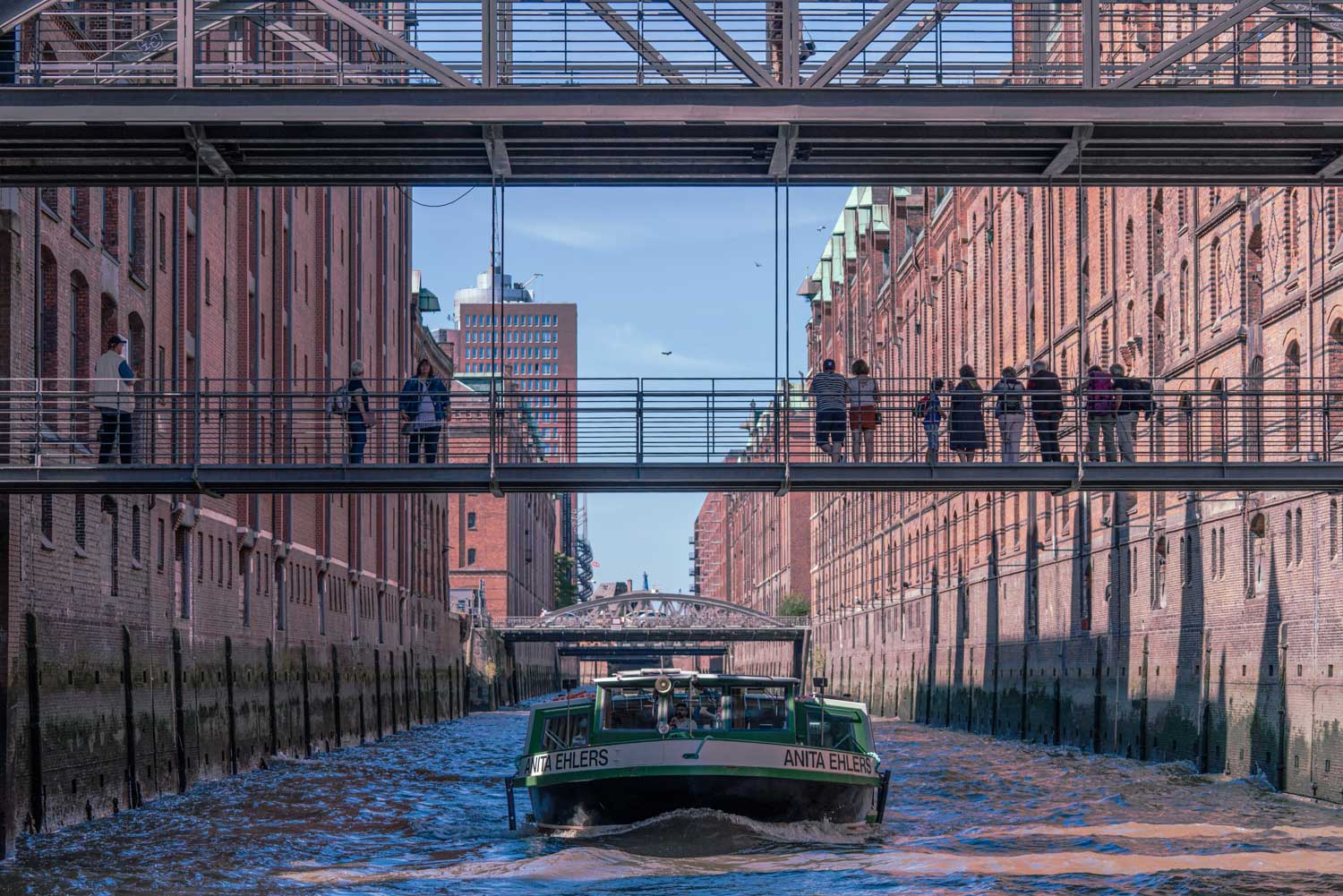 Boat cruising through canal under pedestrian bridges with people in a historic brick district, clear blue sky above.