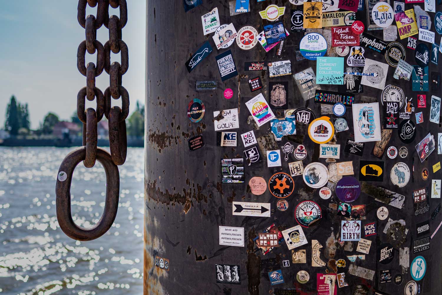 Rusty chain links hang beside a sticker-covered pole by a shimmering river under a bright sky.