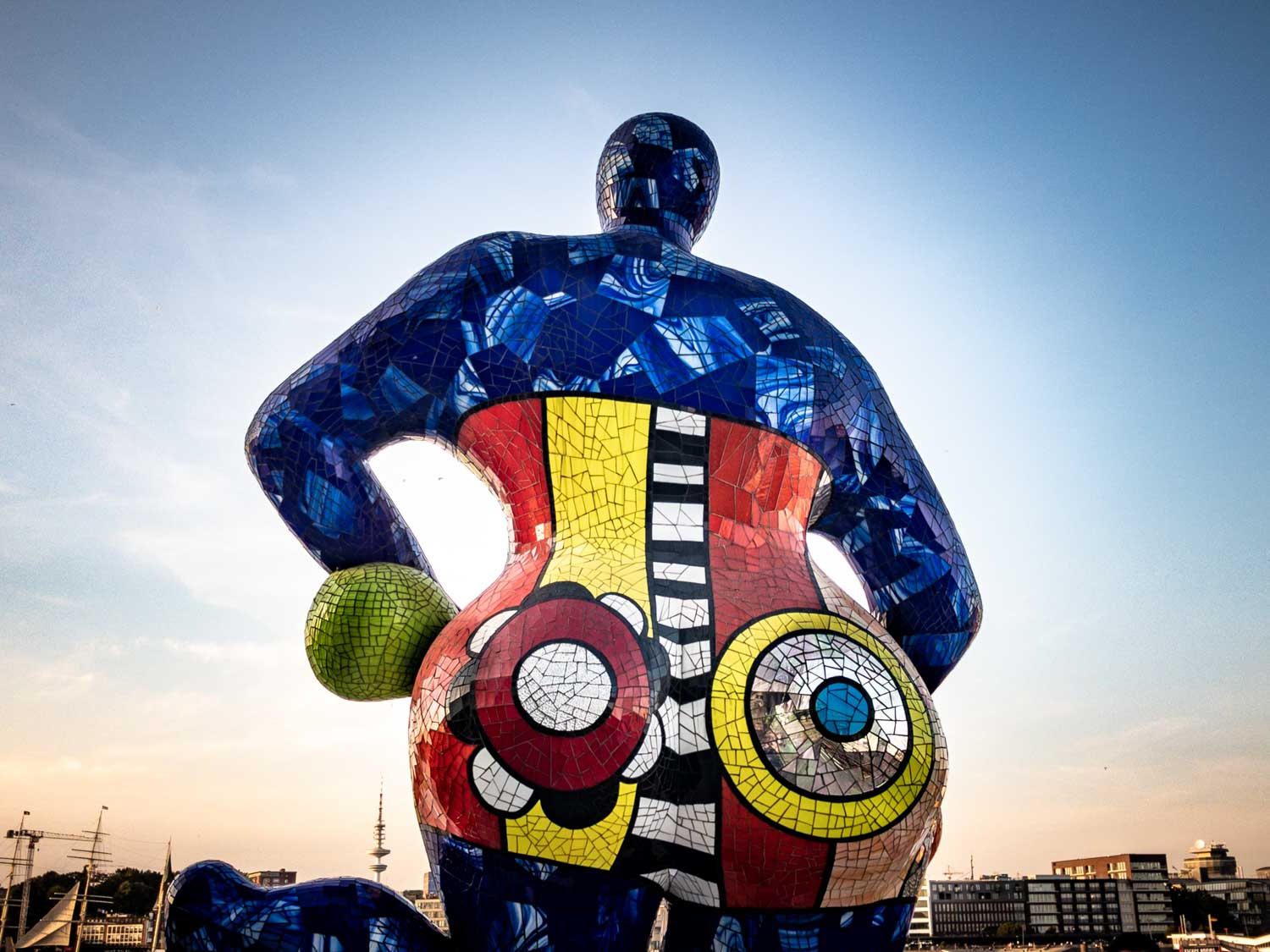 Colorful mosaic sculpture of a figure with vibrant patterns and a cityscape backdrop under a clear blue sky.