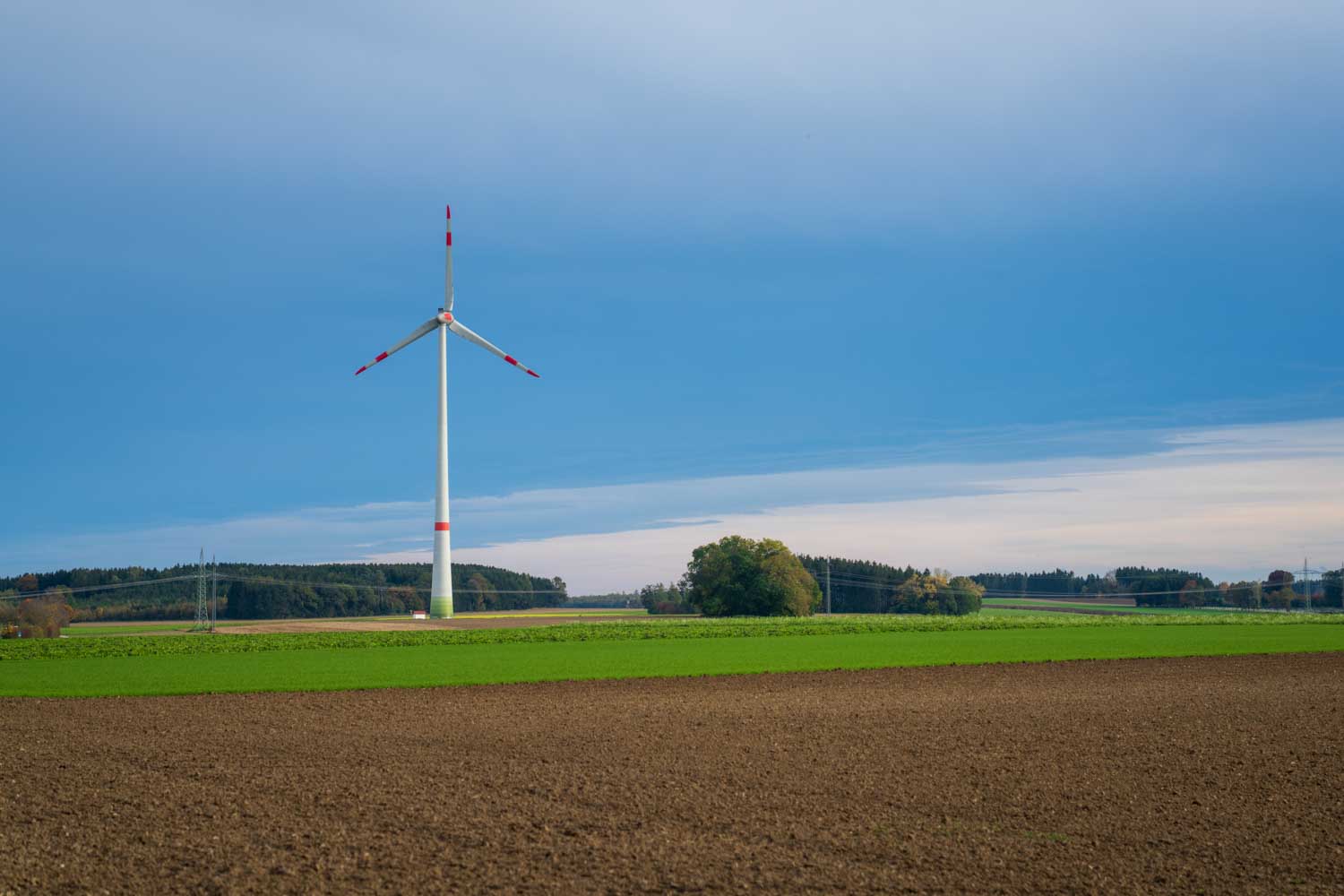 Wind turbine on green field under blue sky, surrounded by farmland and distant forests.