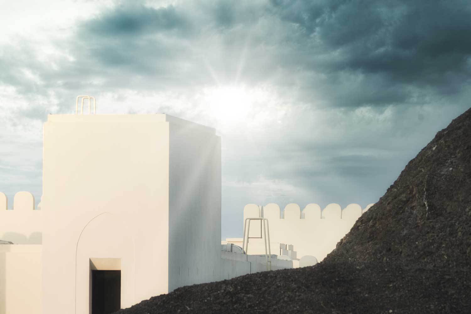 White building with arches under cloudy sky and sun, next to dark hill.