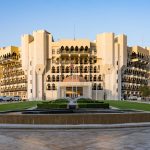 Grand beige building with arched balconies, central entrance, and surrounding cars under a clear blue sky.