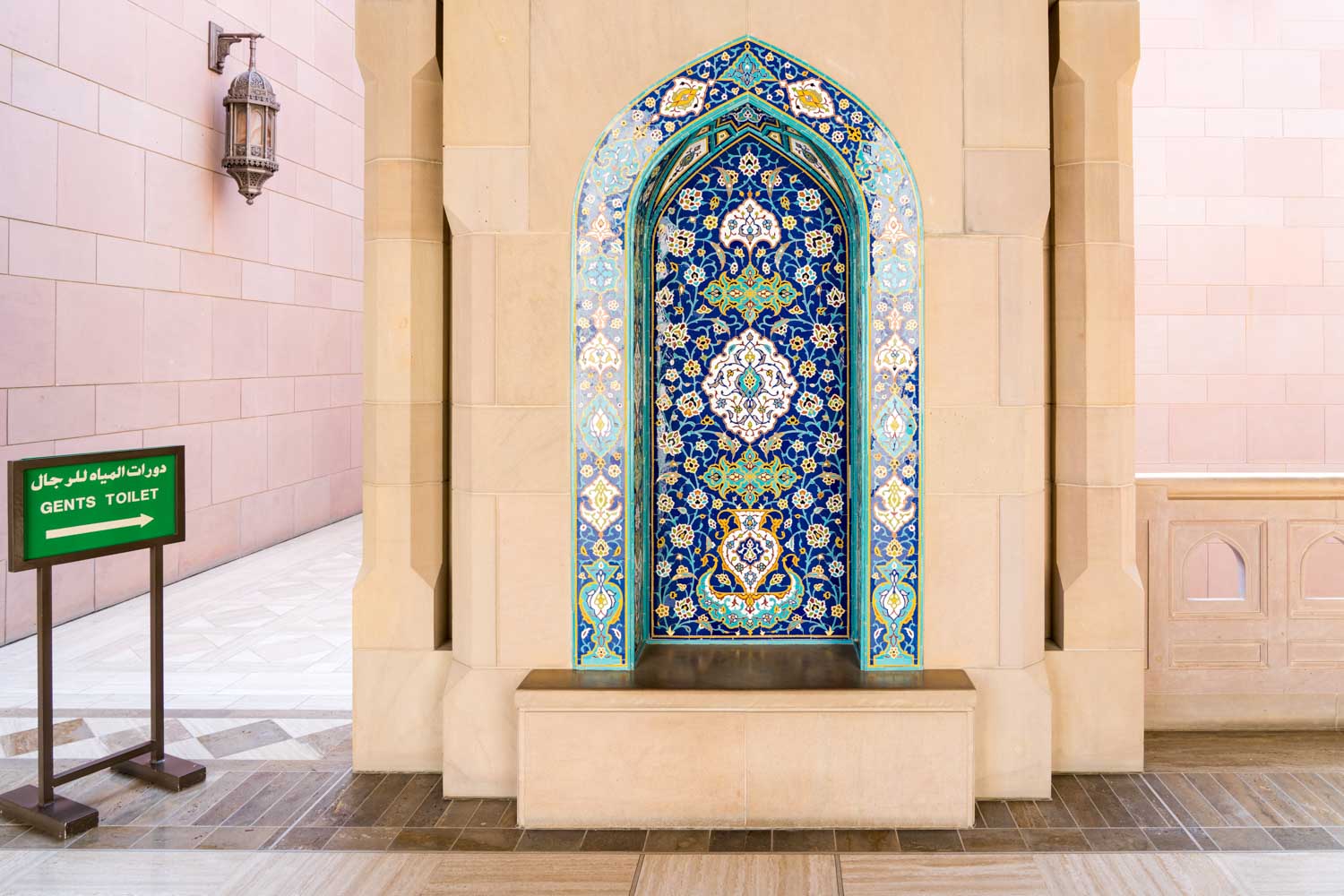 Ornate blue and gold mosaic fountain with Islamic patterns, beside a directional sign for the gents' toilet.