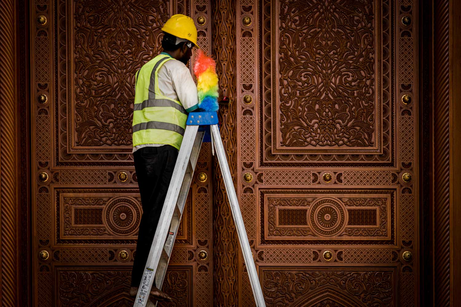 Worker in safety gear dusts ornate wooden door with intricate carvings while standing on a ladder.