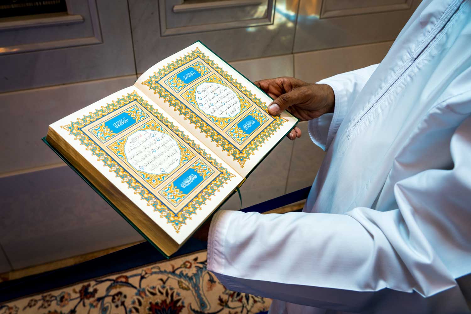 Person in white robe holding an open Quran with ornate Arabic script and colorful designs indoors.