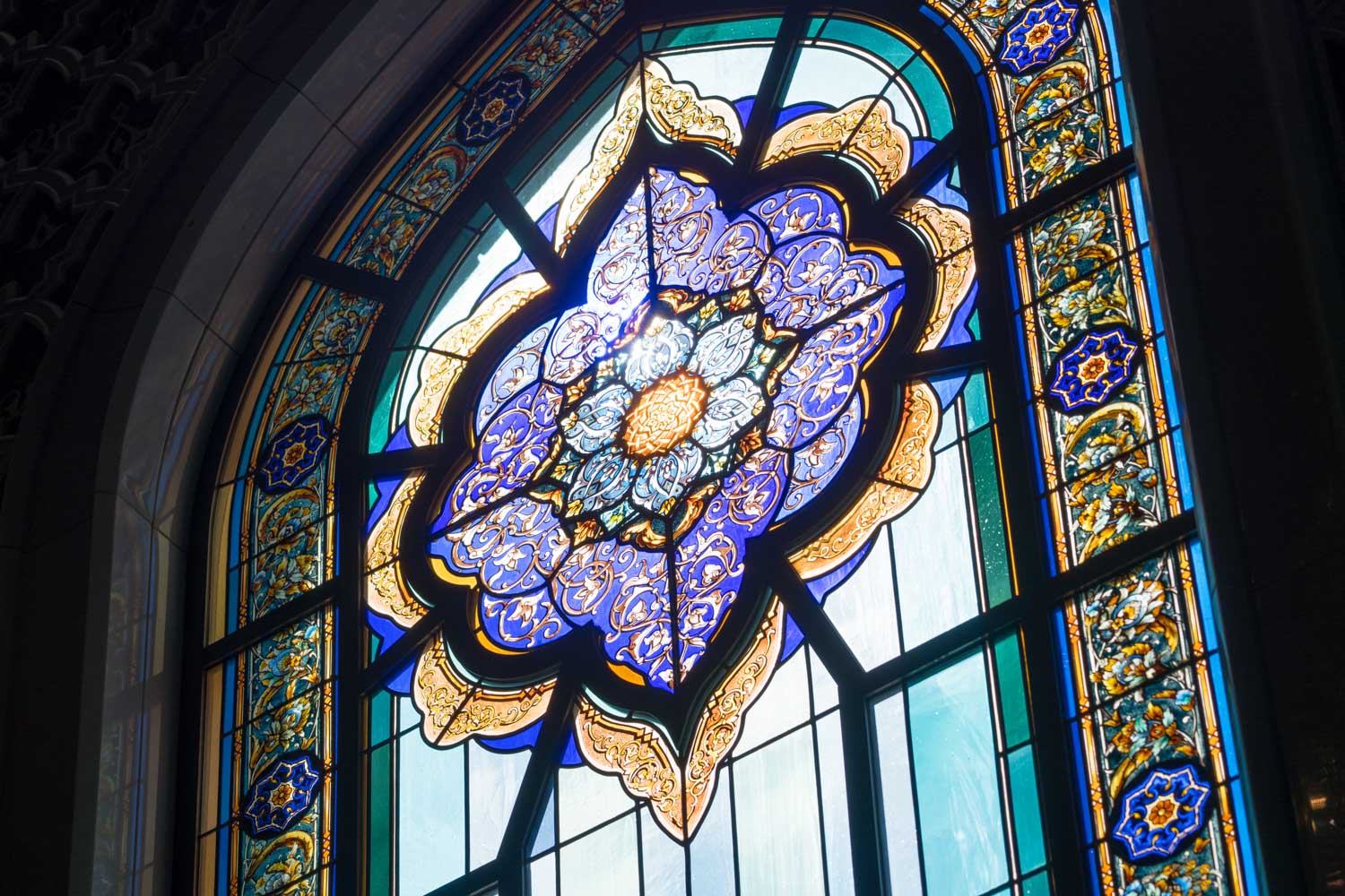 Ornate stained glass window with intricate blue and gold floral patterns, illuminated by natural light.