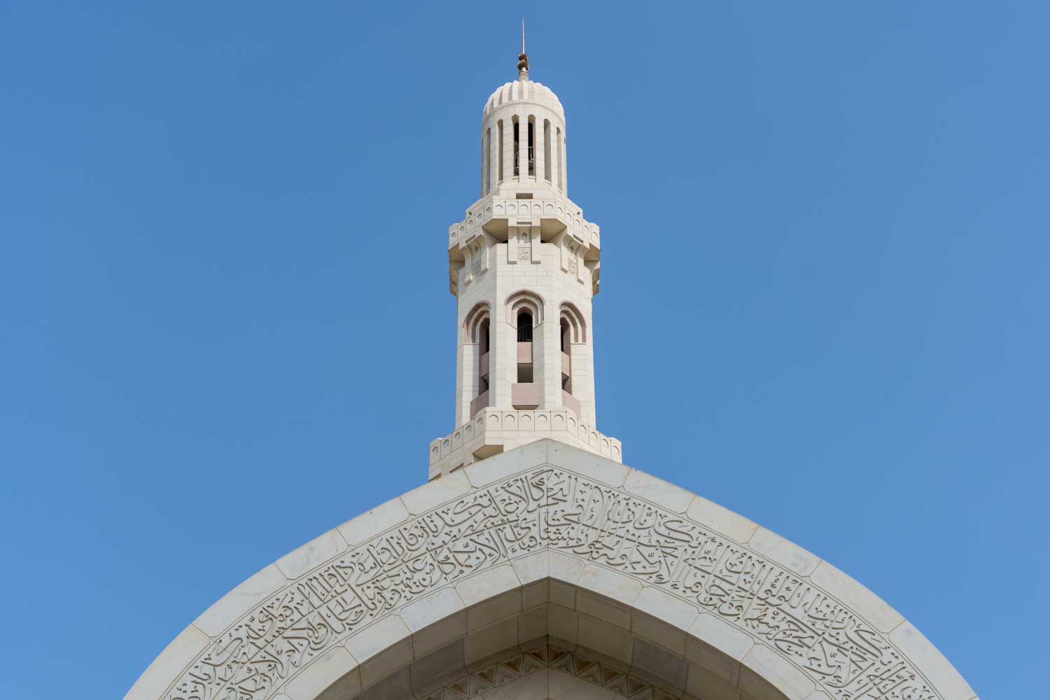 White mosque minaret with intricate Islamic calligraphy on arch set against a clear blue sky.