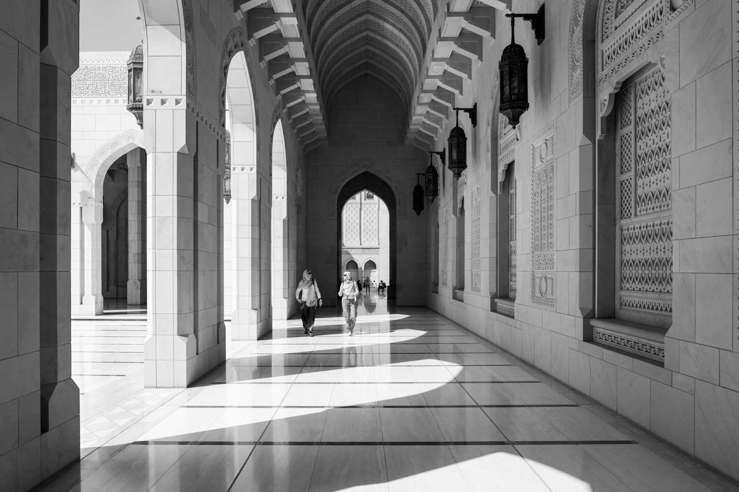 People walking through ornate marble archway in a mosque corridor with intricate wall designs and hanging lanterns.