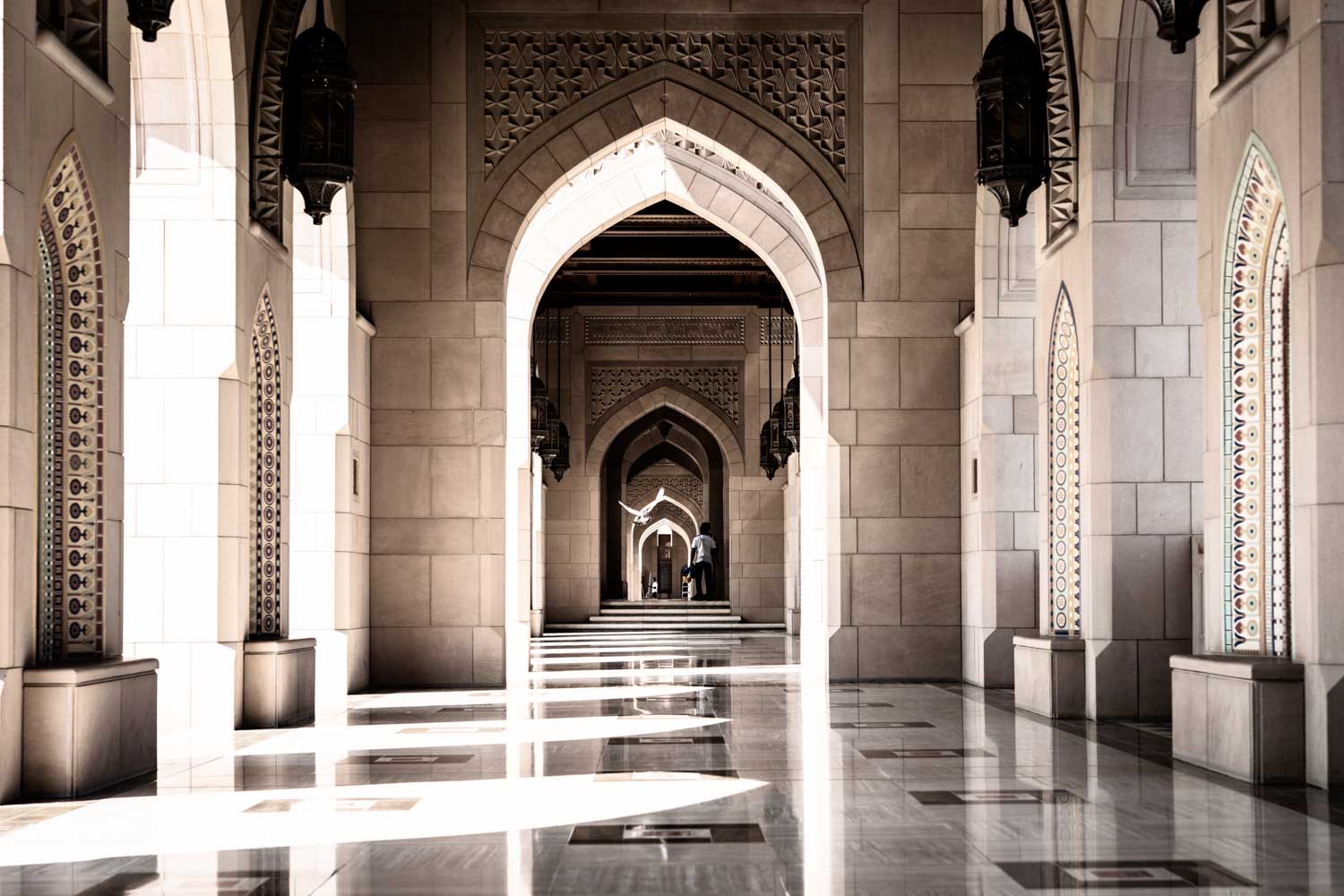 Ornate stone archways in a sunlit corridor with intricate patterns and shadow play on the polished floor.