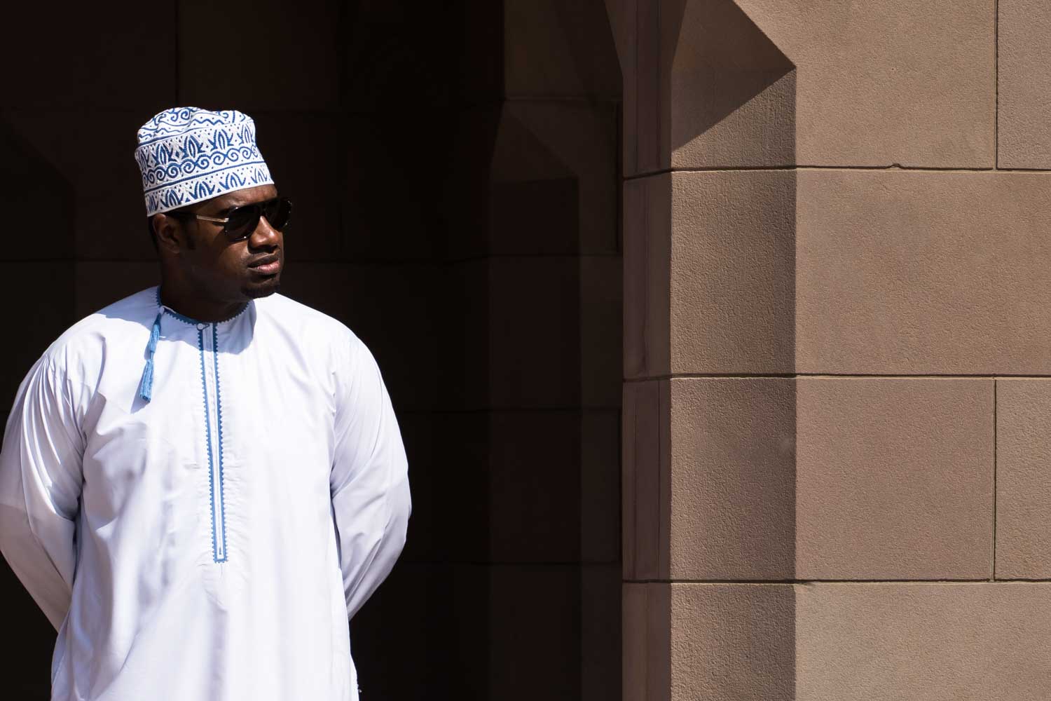 Man in white thobe and kufi stands beside a stone wall, casting a shadow in sunlight.
