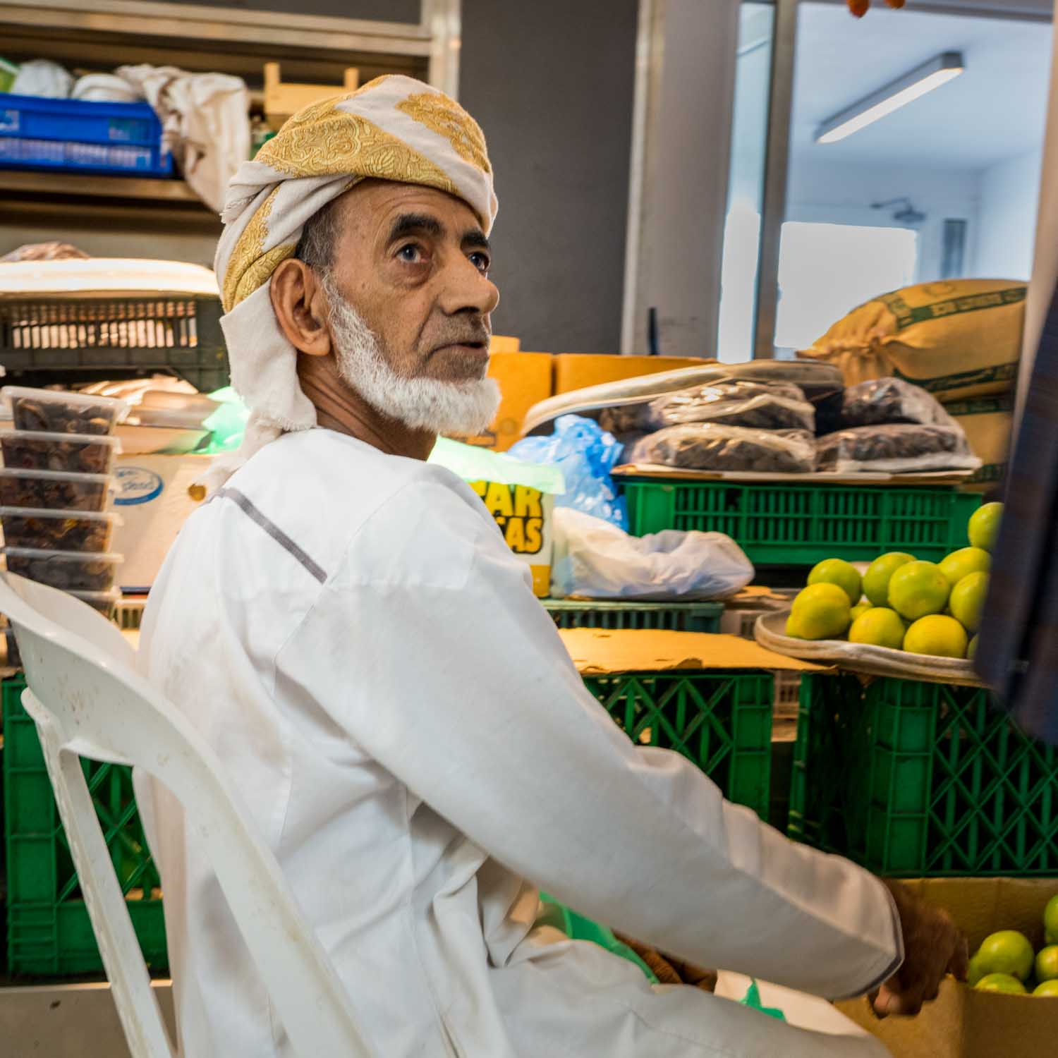 A man in traditional attire sits near crates of fruit at a market stall, surrounded by produce and packaged goods.