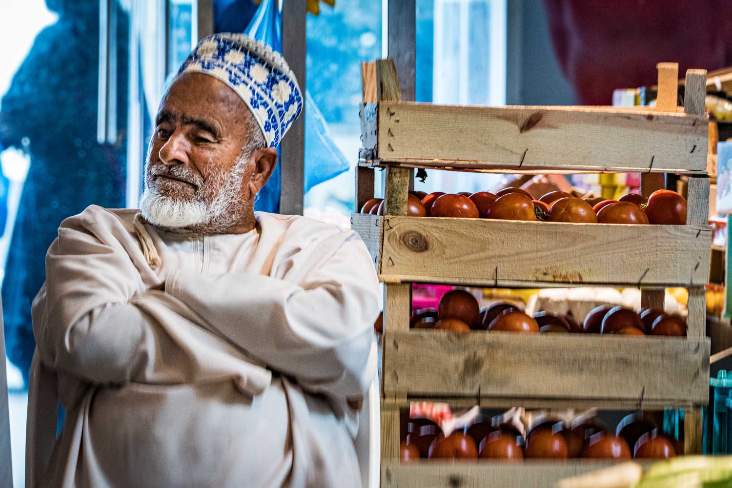 Elderly man in traditional attire sitting by crates of fresh tomatoes in a market setting.