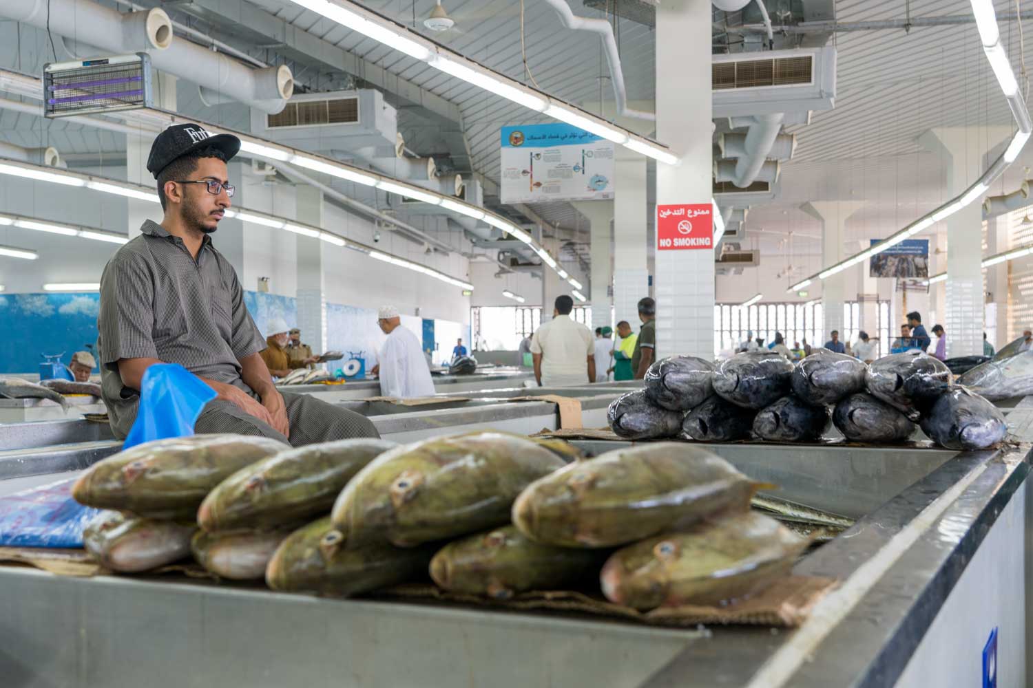 Fish market interior with fresh fish displayed on counters and a seated vendor, surrounded by shoppers under bright lighting.