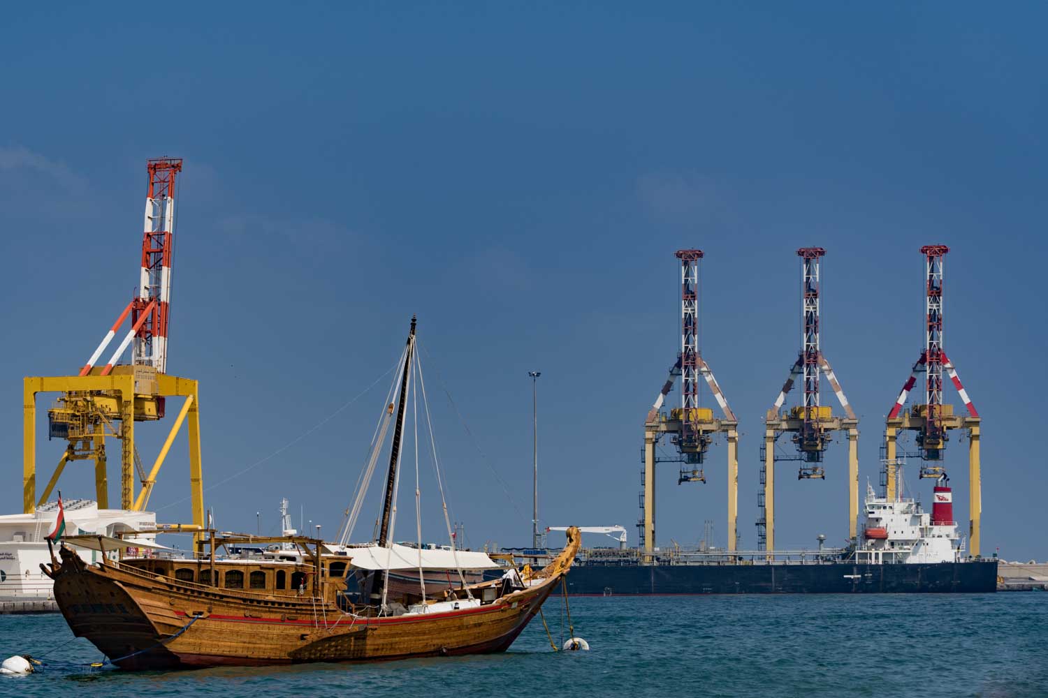 Traditional dhow boat in harbor with large cranes and cargo ship in the background under a clear blue sky.