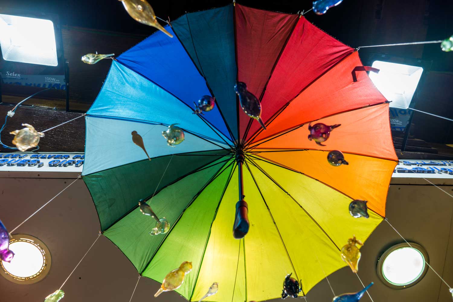 Colorful umbrella with rainbow segments and suspended glass birds, viewed from below with lights in the background.